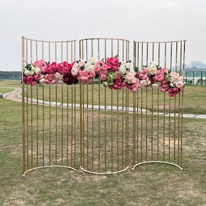 May include: A gold metal backdrop with a curved design, decorated with pink and white flowers. The backdrop is made of thin metal bars and is perfect for creating a beautiful and elegant background for photos or events.