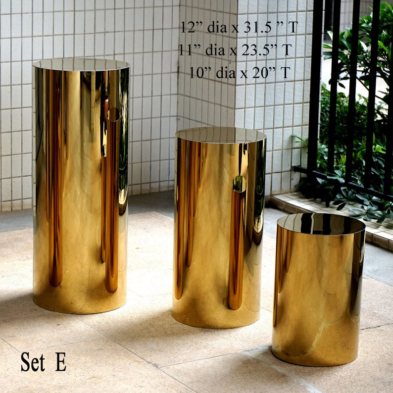 Puede incluir: Conjunto de tres pedestales cil&iacute;ndricos dorados. El m&aacute;s grande tiene 30.5 cm de di&aacute;metro y 80 cm de alto. El del medio tiene 28 cm de di&aacute;metro y 60 cm de alto. El m&aacute;s peque&ntilde;o tiene 25.5 cm de di&aacute;metro y 51 cm de alto. El texto "Set E" est&aacute; abajo.