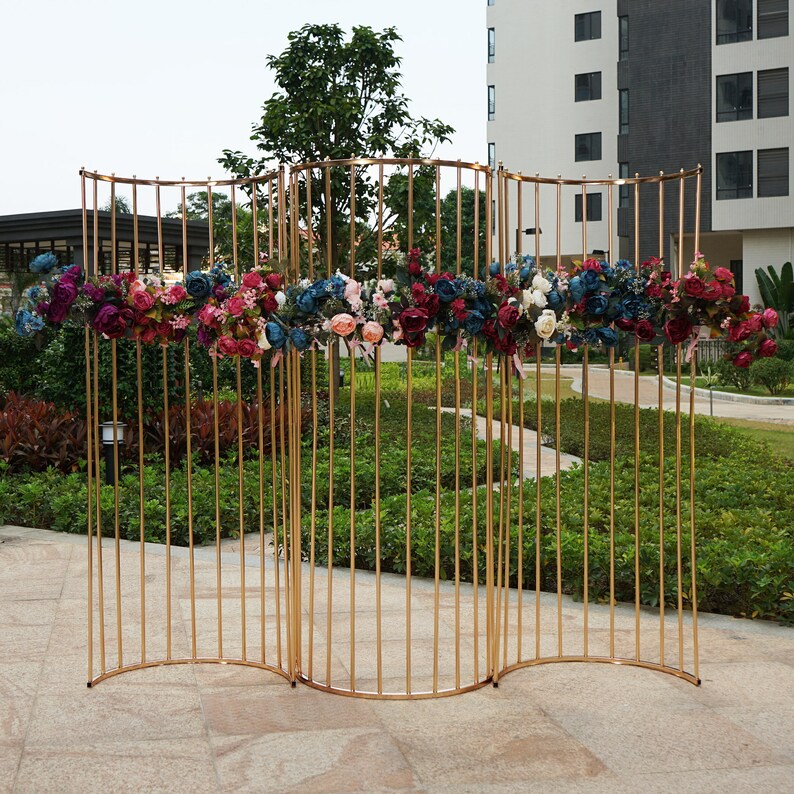 May include: A gold metal backdrop with a curved design, decorated with a garland of burgundy, pink, and blue flowers. The backdrop is made of thin metal rods and is freestanding.