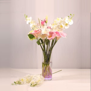 May include: A bouquet of pink and white calla lilies and orchids in a clear glass vase with a purple light at the bottom.