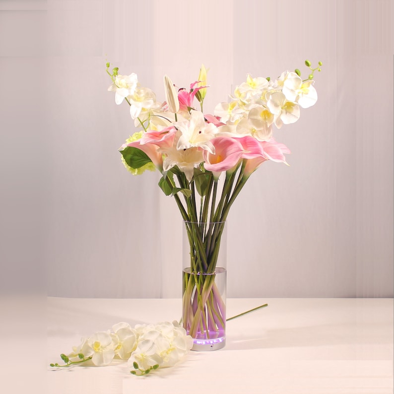 May include: A bouquet of pink and white calla lilies and orchids in a clear glass vase with a purple light at the bottom.