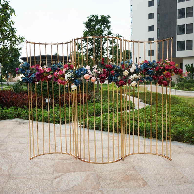 May include: A gold metal backdrop with a curved design, decorated with a garland of pink, red, and blue flowers. The backdrop is made of thin metal rods and is freestanding.