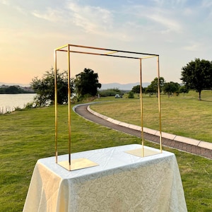 Può includere: Un arco in metallo dorato con una base quadrata, in piedi su una tovaglia bianca con un motivo floreale. L'arco si trova in un ambiente esterno erboso.