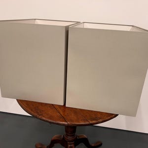 May include: Two light grey square lampshades sitting on a wooden table.
