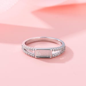 May include: A silver-toned ring featuring a central rectangular flat setting flanked by two split bands encrusted with small clear stones. The design includes a smooth metal surface in the middle, surrounded by a pavé-style setting on both sides.