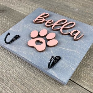 K&ouml;nnte beinhalten: Ein grauer Holz-Hunde-Leinenhalter mit zwei schwarzen Haken. Der Halter hat einen rosa Pfotenabdruck mit einem Herz in der Mitte und den Namen "Bella" in braunen Buchstaben.