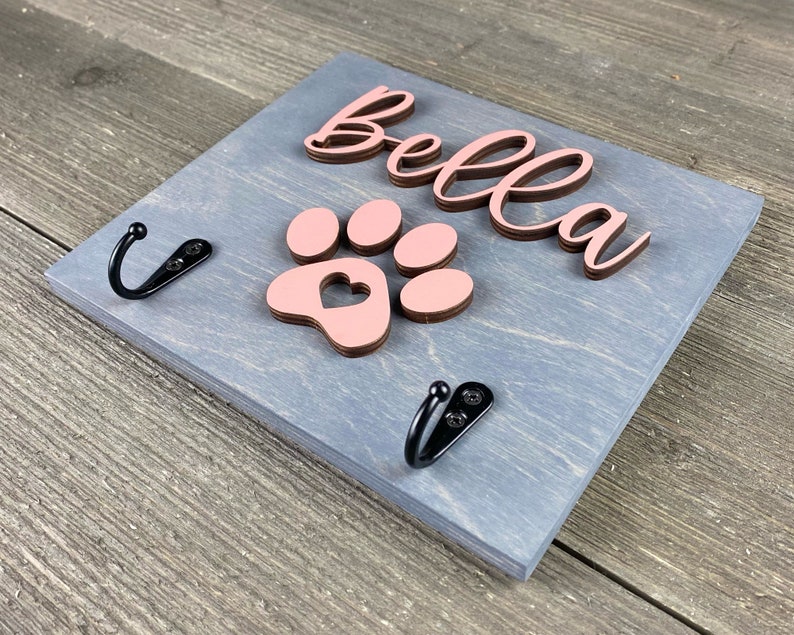 K&ouml;nnte beinhalten: Ein grauer Holz-Hunde-Leinenhalter mit zwei schwarzen Haken. Der Halter hat einen rosa Pfotenabdruck mit einem Herz in der Mitte und den Namen "Bella" in braunen Buchstaben.