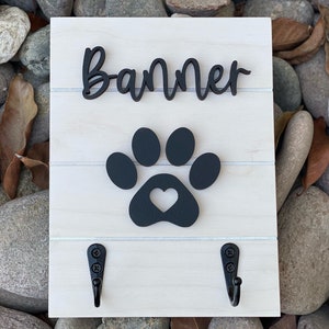 K&ouml;nnte beinhalten: Ein wei&szlig;es Holzschild mit dem Namen "Banner" in schwarzen Kursivbuchstaben, einem schwarzen Pfotenabdruck mit einem Herz in der Mitte und zwei schwarzen Haken.