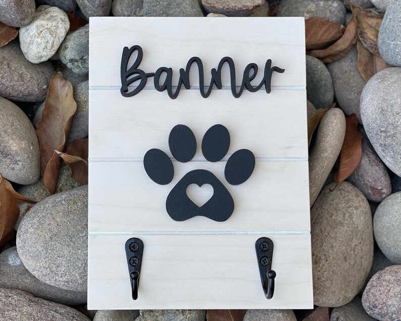 K&ouml;nnte beinhalten: Ein wei&szlig;es Holzschild mit dem Namen "Banner" in schwarzen Kursivbuchstaben, einem schwarzen Pfotenabdruck mit einem Herz in der Mitte und zwei schwarzen Haken.