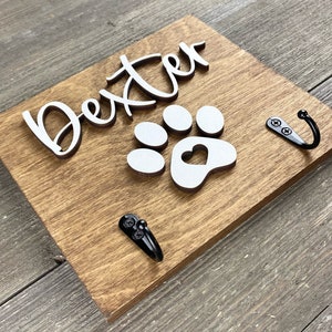 K&ouml;nnte beinhalten: Ein Holzschild mit dem Namen "Dexter" in wei&szlig;en Buchstaben und einem wei&szlig;en Pfotenabdruck mit einem Herz in der Mitte. Das Schild hat zwei schwarze Metallhaken.