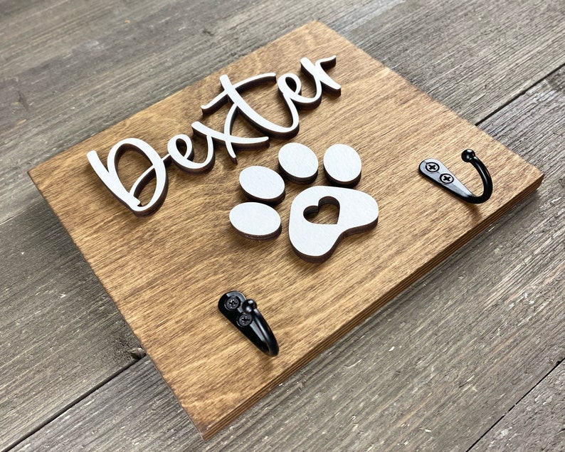 K&ouml;nnte beinhalten: Ein Holzschild mit dem Namen "Dexter" in wei&szlig;en Buchstaben und einem wei&szlig;en Pfotenabdruck mit einem Herz in der Mitte. Das Schild hat zwei schwarze Metallhaken.