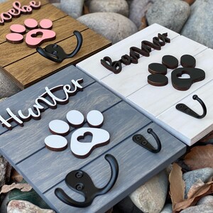 K&ouml;nnte beinhalten: Drei Haken aus Holz f&uuml;r Hundeleinen mit Pfotenabdruck-Designs und personalisierten Namen. Die Haken sind in verschiedenen Farben lackiert: Braun, Wei&szlig; und Grau. Die Namen auf den Haken sind "Phoebe", "Bama" und "Hunter".