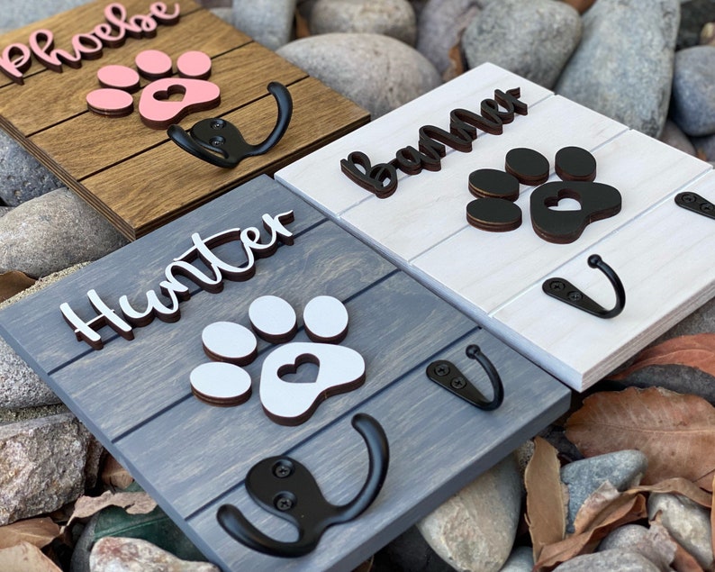 K&ouml;nnte beinhalten: Drei Haken aus Holz f&uuml;r Hundeleinen mit Pfotenabdruck-Designs und personalisierten Namen. Die Haken sind in verschiedenen Farben lackiert: Braun, Wei&szlig; und Grau. Die Namen auf den Haken sind "Phoebe", "Bama" und "Hunter".