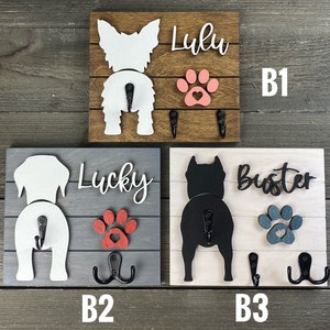 May include: Three wooden dog leash holders with paw prints and dog silhouettes. The top holder is brown with a white dog silhouette and the name "Lulu". The middle holder is gray with a white dog silhouette and the name "Lucky". The bottom holder is white with a black dog silhouette and the name "Buster".