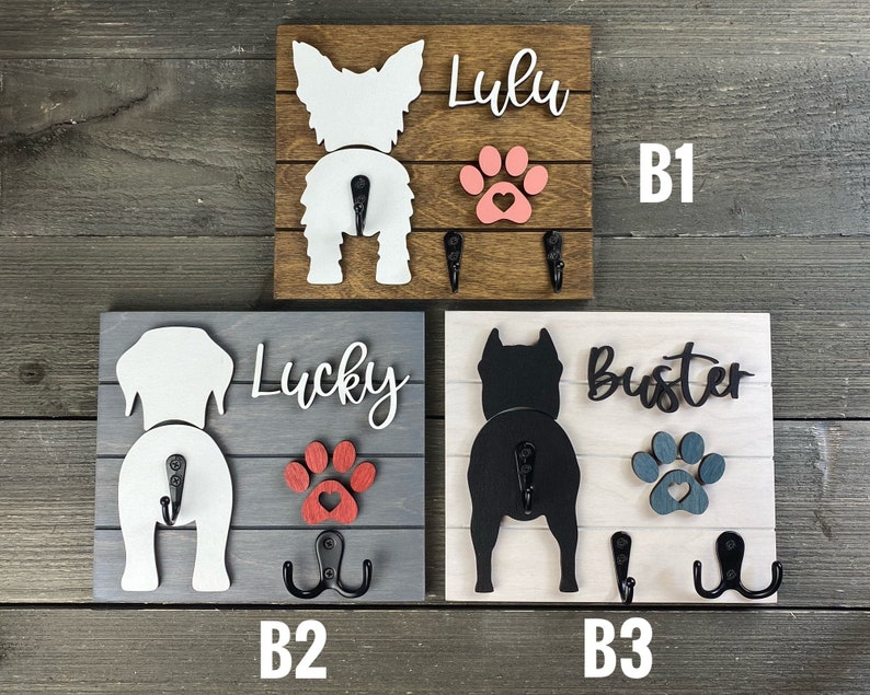 May include: Three wooden dog leash holders with paw prints and dog silhouettes. The top holder is brown with a white dog silhouette and the name "Lulu". The middle holder is gray with a white dog silhouette and the name "Lucky". The bottom holder is white with a black dog silhouette and the name "Buster".