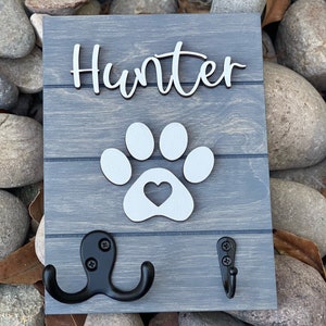 K&ouml;nnte beinhalten: Ein graues Holzschild mit dem Namen "Hunter" in wei&szlig;er Kursivschrift. Unter dem Namen befindet sich ein wei&szlig;er Pfotenabdruck mit einem Herz in der Mitte. Zwei schwarze Metallhaken sind an der Unterseite des Schildes befestigt.