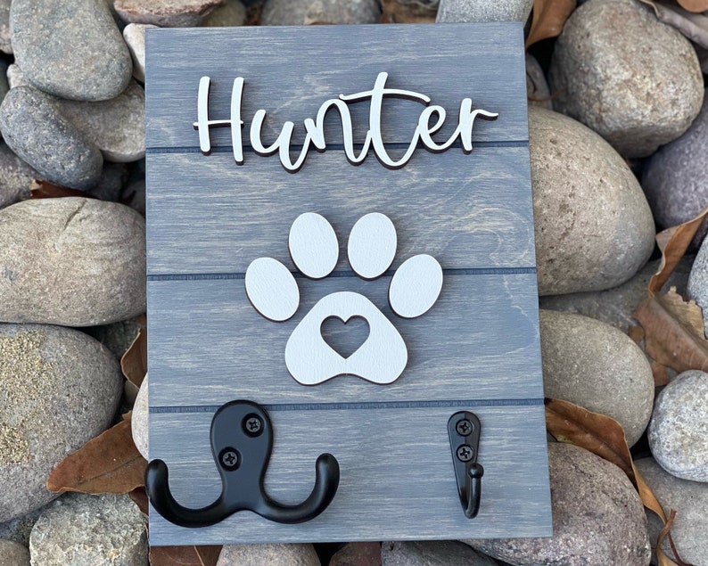 K&ouml;nnte beinhalten: Ein graues Holzschild mit dem Namen "Hunter" in wei&szlig;er Kursivschrift. Unter dem Namen befindet sich ein wei&szlig;er Pfotenabdruck mit einem Herz in der Mitte. Zwei schwarze Metallhaken sind an der Unterseite des Schildes befestigt.