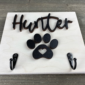 K&ouml;nnte beinhalten: Ein wei&szlig;es Holzschild mit dem Namen "Hunter" in schwarzen Buchstaben und einem schwarzen Pfotenabdruck mit einem Herz in der Mitte. Zwei schwarze Haken sind an der Unterseite des Schildes befestigt.