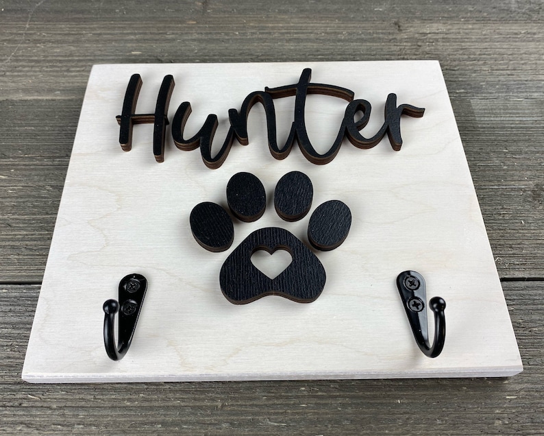 K&ouml;nnte beinhalten: Ein wei&szlig;es Holzschild mit dem Namen "Hunter" in schwarzen Buchstaben und einem schwarzen Pfotenabdruck mit einem Herz in der Mitte. Zwei schwarze Haken sind an der Unterseite des Schildes befestigt.