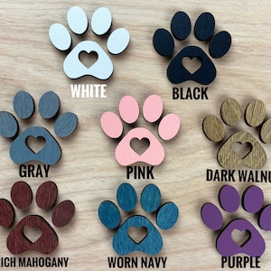 K&ouml;nnte beinhalten: Neun Holz-Pfotenabdr&uuml;cke mit einem Herz-Ausschnitt in der Mitte, jeder in einer anderen Farbe bemalt: wei&szlig;, schwarz, grau, rosa, dunkelbraun, mahagonifarben, abgenutzt marineblau und lila. Die Pfotenabdr&uuml;cke sind in drei Reihen zu je drei angeordnet.