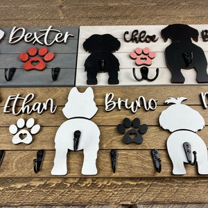 May include: Wooden dog leash rack with four hooks. The rack features four dog silhouettes with paw prints and names: Dexter, Chloe, Bruno, and Max. The rack is painted in shades of gray, white, and brown.