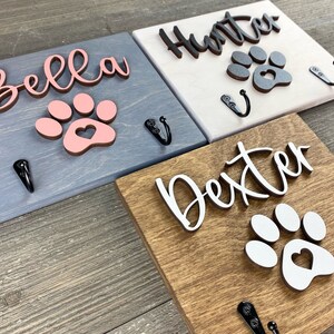 K&ouml;nnte beinhalten: Drei Holz-Haken f&uuml;r Hundeleinen mit Pfotenabdruck-Designs. Die Haken sind in verschiedenen Farben lackiert: blau, wei&szlig; und braun. Die Namen "Bella", "Hunter" und "Dexter" sind auf den Haken geschrieben.