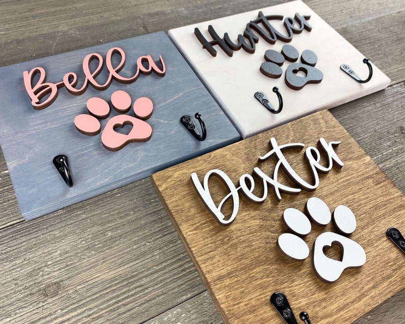 K&ouml;nnte beinhalten: Drei Holz-Haken f&uuml;r Hundeleinen mit Pfotenabdruck-Designs. Die Haken sind in verschiedenen Farben lackiert: blau, wei&szlig; und braun. Die Namen "Bella", "Hunter" und "Dexter" sind auf den Haken geschrieben.