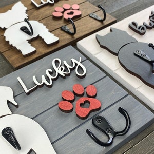 May include: Four wooden dog leash hooks with paw prints and dog silhouettes. The hooks are painted in different colors, including white, black, red, and brown. The hooks are labeled with the names "Lucky", "Buster", and "Lola".