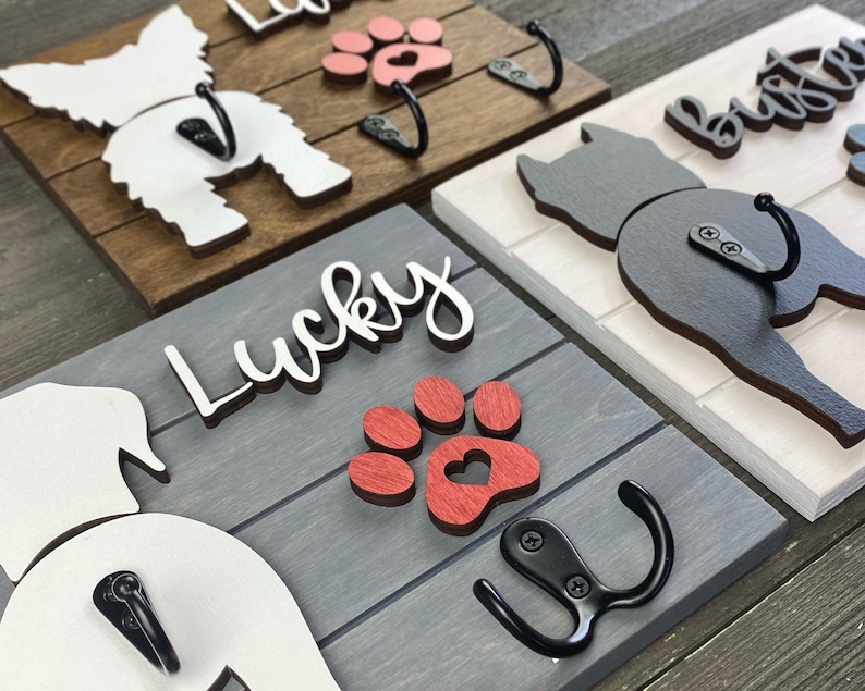 May include: Four wooden dog leash hooks with paw prints and dog silhouettes. The hooks are painted in different colors, including white, black, red, and brown. The hooks are labeled with the names "Lucky", "Buster", and "Lola".