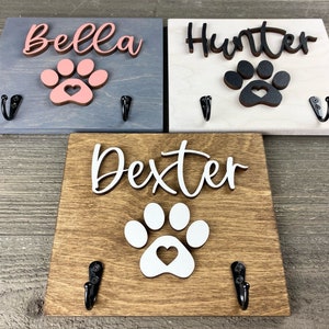 K&ouml;nnte beinhalten: Drei Holz-Haken f&uuml;r Hundeleinen mit Pfotenabdr&uuml;cken und personalisierten Namen: Bella, Hunter und Dexter. Jeder Haken hat zwei Metallhaken zum Aufh&auml;ngen von Leinen.