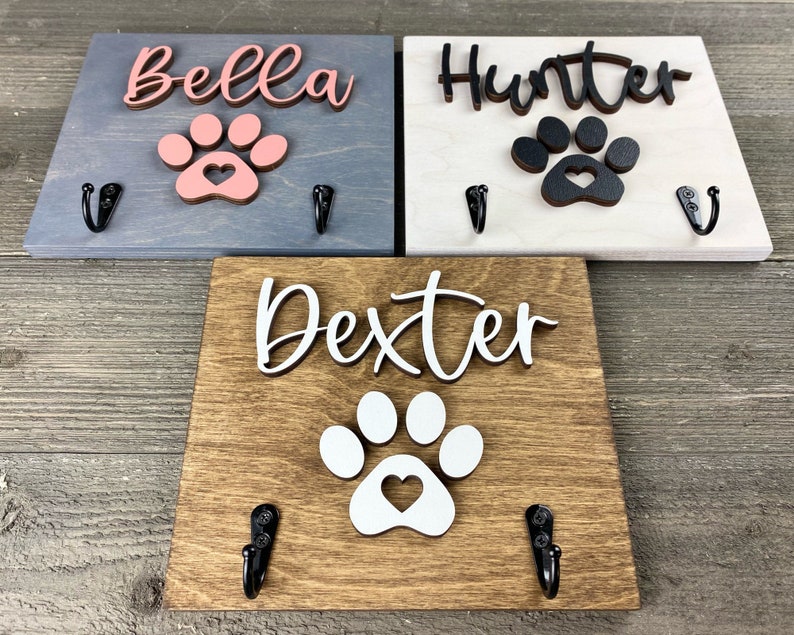 K&ouml;nnte beinhalten: Drei Holz-Haken f&uuml;r Hundeleinen mit Pfotenabdr&uuml;cken und personalisierten Namen: Bella, Hunter und Dexter. Jeder Haken hat zwei Metallhaken zum Aufh&auml;ngen von Leinen.
