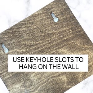 K&ouml;nnte beinhalten: Ein dunkelbraunes Holzschild mit zwei Schl&uuml;ssell&ouml;chern zum Aufh&auml;ngen an einer Wand. Das Schild tr&auml;gt die Aufschrift "USE KEYHOLE SLOTS TO HANG ON THE WALL".