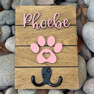 K&ouml;nnte beinhalten: Haken aus Holz f&uuml;r die Hundeleine mit einem rosa Pfotenabdruck und dem Namen "Phoebe" in rosa kursiver Schrift.
