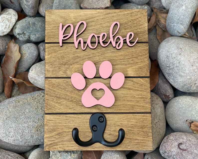 K&ouml;nnte beinhalten: Haken aus Holz f&uuml;r die Hundeleine mit einem rosa Pfotenabdruck und dem Namen "Phoebe" in rosa kursiver Schrift.