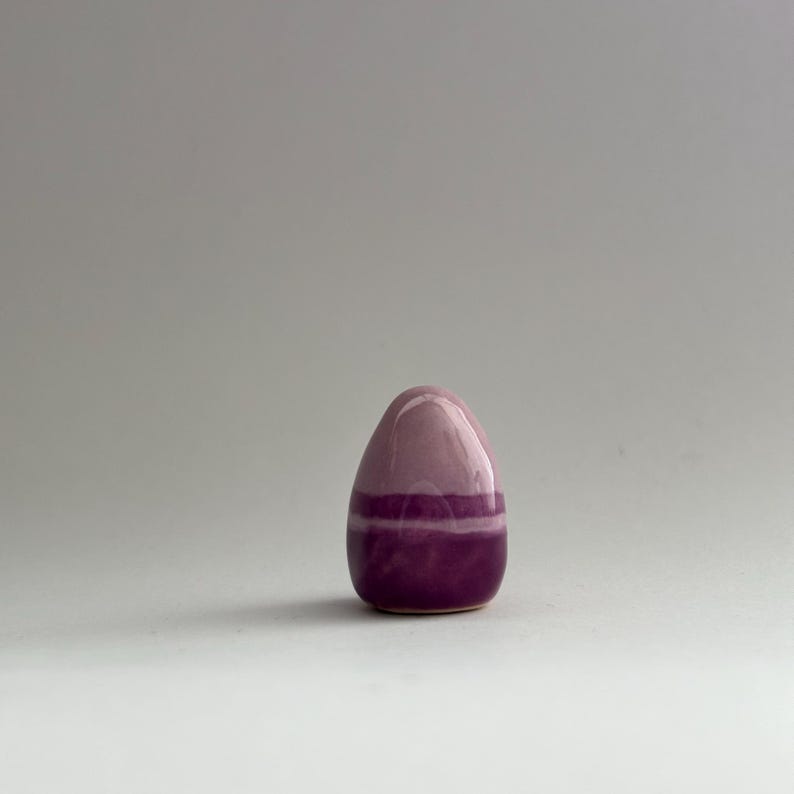 K&ouml;nnte beinhalten: Ein kleines, eif&ouml;rmiges Objekt aus Keramik mit einer rosa und violetten Glasur. Das Ei hat eine glatte, gl&auml;nzende Oberfl&auml;che und ist mit einem d&uuml;nnen, horizontalen Streifen in Violett verziert.