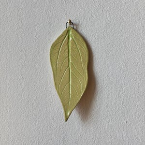 Puede incluir: Un colgante en forma de hoja verde claro con un patrón de vetas detallado. El colgante está suspendido de un pequeño lazo dorado y un alambre negro. La hoja tiene un acabado brillante.