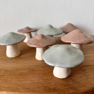 Peut inclure: Plusieurs figurines de champignons en céramique aux couleurs douces, notamment gris, marron et crème. Les champignons ont des chapeaux arrondis et des surfaces lisses et brillantes, disposés sur une surface en bois.