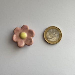 May include: A small pink ceramic flower with a yellow centre. The flower is about the size of a UK 20 pence coin.