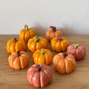 Op de afbeelding: Een verzameling keramische pompoenen in verschillende tinten oranje en roze, met bruine en groene stelen. De pompoenen zijn gerangschikt op een houten oppervlak, perfect voor herfstdecoratie.