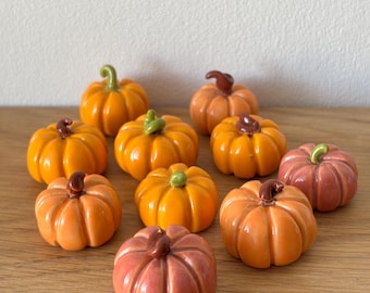 May include: A collection of ceramic pumpkins in various shades of orange and pink, with brown and green stems. The pumpkins are arranged on a wooden surface, perfect for fall decor.