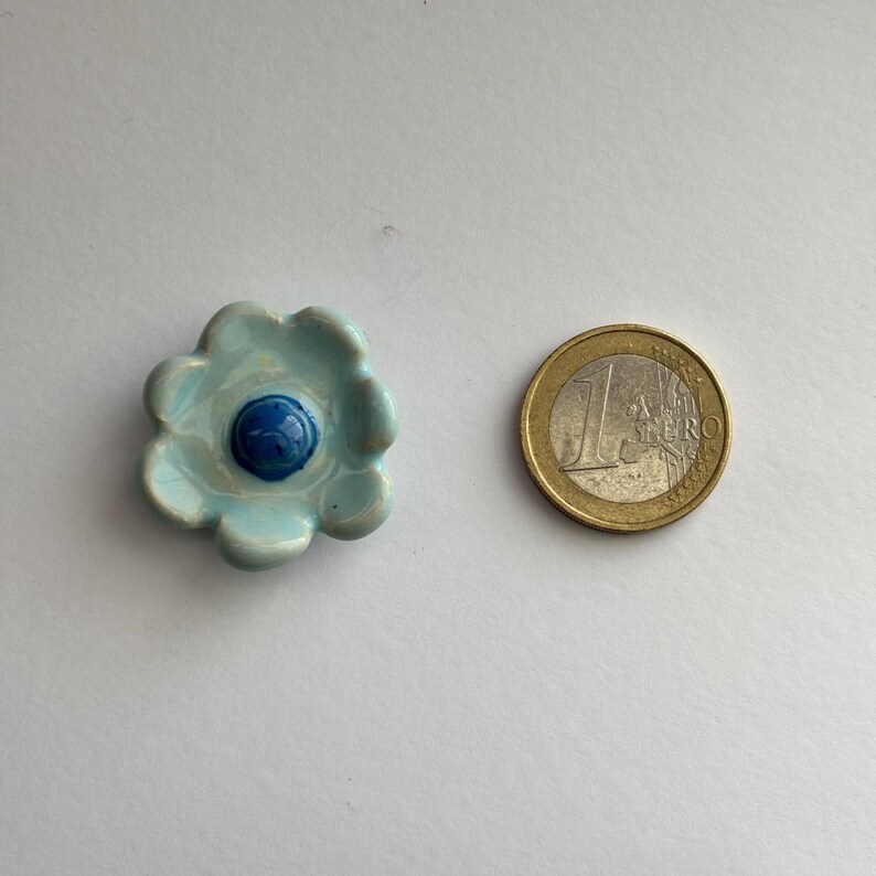 May include: A light blue ceramic flower with a blue centre. The flower is about the size of a euro coin.