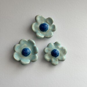 May include: Three ceramic flower-shaped buttons with a blue centre. The flowers are light blue with a slightly iridescent finish.