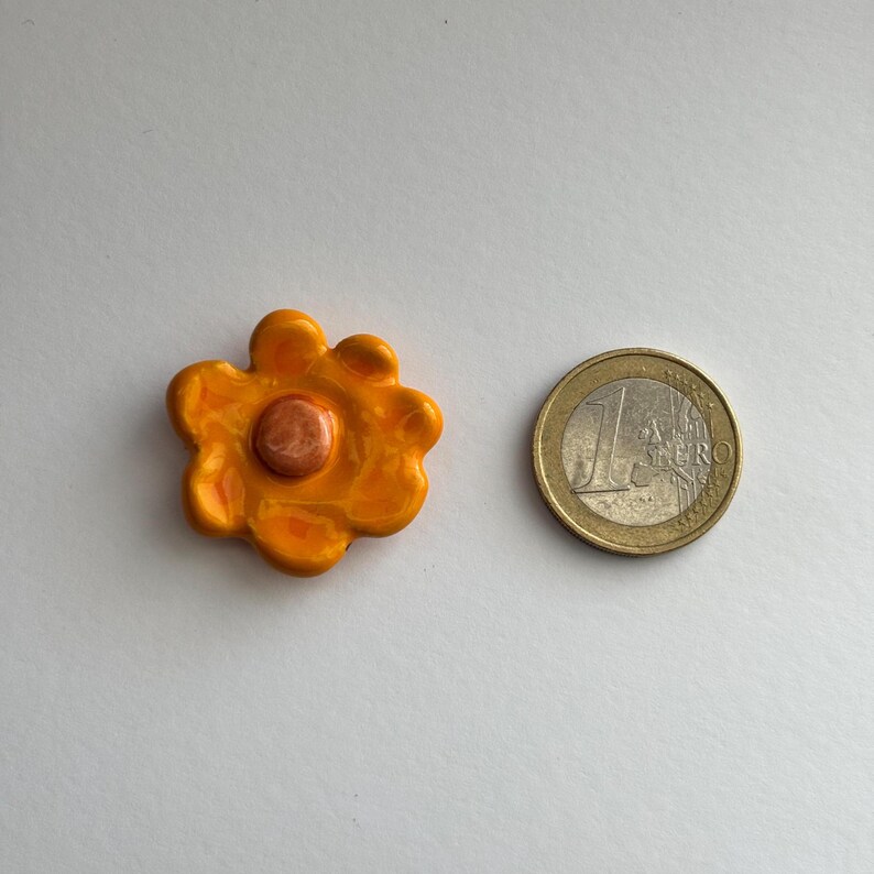 May include: A small, orange ceramic flower with a raised, orange centre. The flower is about the size of a UK one euro coin.