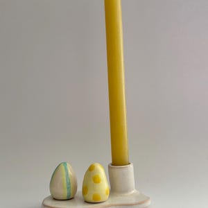 K&ouml;nnte beinhalten: Wei&szlig;er Kerzenhalter aus Keramik mit zwei kleinen eif&ouml;rmigen Haltern. Ein Ei ist mit gelben Punkten verziert, das andere mit gr&uuml;nen und gelben Streifen. Eine gelbe Kerze befindet sich im mittleren Halter.