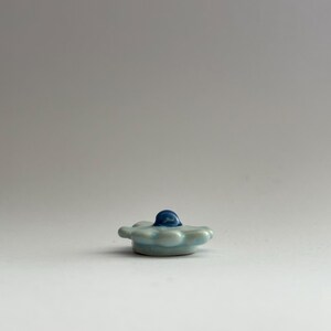 May include: A small, light blue ceramic flower with a blue centre. The flower has a rounded shape and is slightly raised in the centre.