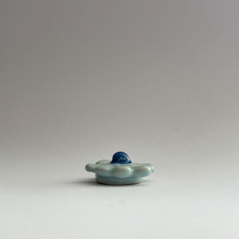 May include: A small, light blue ceramic flower with a blue centre. The flower has a rounded shape and is slightly raised in the centre.
