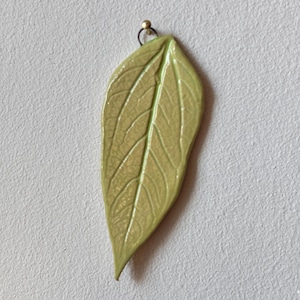 Op de afbeelding: Een groene keramische bladeren hanger met een gouden metalen lus aan de bovenkant.