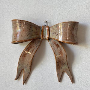 May include: A ceramic bow wall hanging with a speckled, earthy color palette. The bow features a textured surface with brown, tan, and black speckles. The bow's design includes two loops and two flowing tails, with a small metal loop for hanging.
