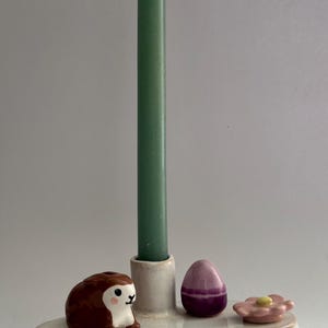 K&ouml;nnte beinhalten: Ein wei&szlig;er Kerzenhalter aus Keramik mit einer gr&uuml;nen Kerze, einer braunen Hasenfigur, einem lila Ei und einer rosa Blume.