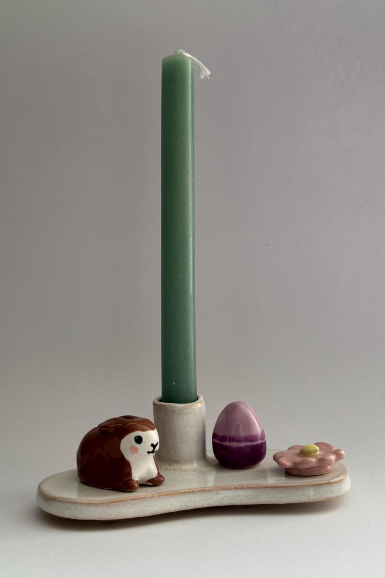 K&ouml;nnte beinhalten: Ein wei&szlig;er Kerzenhalter aus Keramik mit einer gr&uuml;nen Kerze, einer braunen Hasenfigur, einem lila Ei und einer rosa Blume.